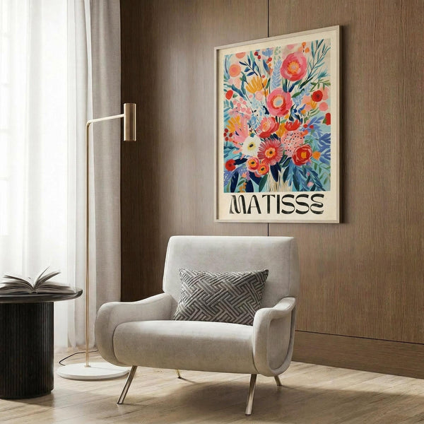 May include: Modern living room with beige armchair and geometric pillow, brass floor lamp, dark side table with open book, and framed floral art print on wood-paneled wall. The colorful botanical artwork features vibrant pink, coral, blue and yellow flowers in an abstract style with "MATISSE" text below the bouquet design.