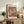 May include: Framed art print featuring abstract cut-out style shapes in warm oranges, reds, and yellows with "MATISSE" and "PAPIERS DÉCOUPÉS" text, displayed on rustic wooden sideboard beside fern plant, books, ceramic vases, and beige upholstered chair in neutral-toned living room setting.