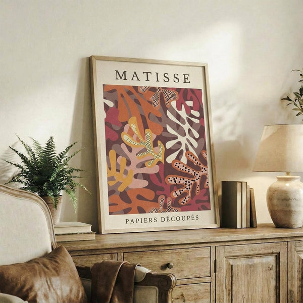 May include: Framed art print featuring abstract cut-out style shapes in warm oranges, reds, and yellows with "MATISSE" and "PAPIERS DÉCOUPÉS" text, displayed on rustic wooden sideboard beside fern plant, books, ceramic vases, and beige upholstered chair in neutral-toned living room setting.