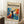 May include: Framed poster leaning against white wall corner featuring colorful abstract figure with raised arms in blues, oranges, and reds. Text reads "matisse modernist" with exhibition details. White coffee cup on wooden block nearby, beige blanket visible, poster sits on textured white rug.