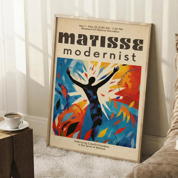 May include: Framed poster leaning against white wall corner featuring colorful abstract figure with raised arms in blues, oranges, and reds. Text reads "matisse modernist" with exhibition details. White coffee cup on wooden block nearby, beige blanket visible, poster sits on textured white rug.