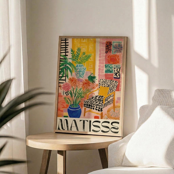 May include: Colorful Matisse-style poster in light wood frame showing interior scene with patterned armchair, potted plants, decorative tiles, and vibrant botanical elements in yellow, coral, green, and blue tones. Displayed on round wooden side table next to beige upholstered chair in bright, naturally-lit room with white walls and plant shadows.