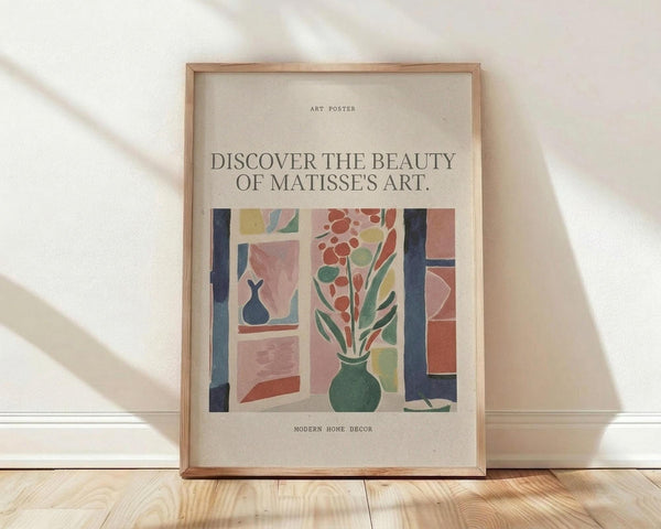 May include: Framed art poster with cream background featuring colorful Matisse-style interior scene showing green vase with red flowers and foliage viewed through window frame with blue curtains and pink geometric shapes. Text reads "Discover the beauty of Matisse's art" and "Modern home decor." Natural wood frame leaning against white wall on light wooden floor with soft window shadows.