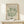 May include: Framed botanical art print in natural wood frame displaying green coral-like forms on cream background, positioned on light wood console table beside small wrapped gift with twine and evergreen sprig against white wall