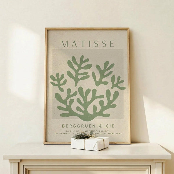 May include: Framed botanical art print in natural wood frame displaying green coral-like forms on cream background, positioned on light wood console table beside small wrapped gift with twine and evergreen sprig against white wall