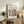 May include: Framed art poster with colorful Matisse-inspired illustration featuring vases with flowers and foliage in coral, teal, and yellow tones, displayed on rustic wooden sideboard alongside fern plant, ceramic vase, stacked books, and cream upholstered chair in neutral-toned living room setting with natural lighting