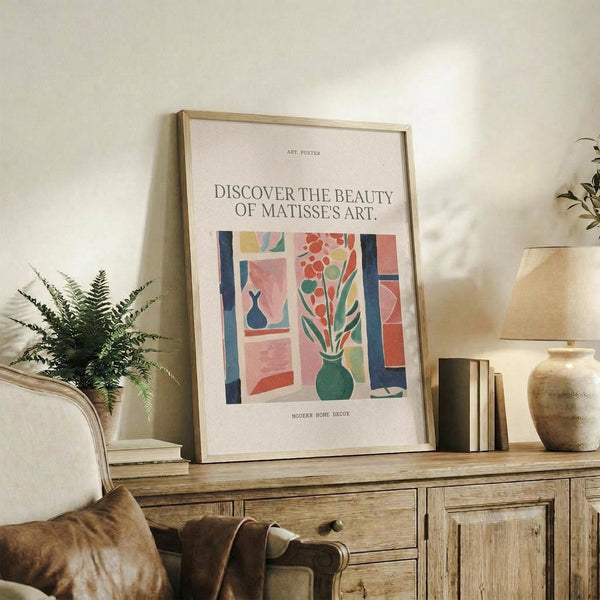May include: Framed art poster with colorful Matisse-inspired illustration featuring vases with flowers and foliage in coral, teal, and yellow tones, displayed on rustic wooden sideboard alongside fern plant, ceramic vase, stacked books, and cream upholstered chair in neutral-toned living room setting with natural lighting