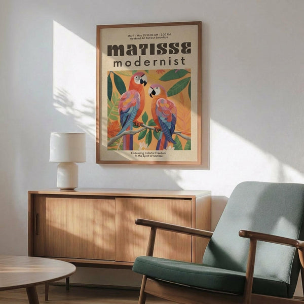 May include: Framed vintage-style poster featuring two colorful parrots perched on branches with tropical leaves, displaying vibrant reds, blues, and greens against a warm yellow background. The poster hangs above a mid-century modern wooden sideboard with a white ceramic table lamp, next to a sage green upholstered armchair with wooden arms in a sunlit room with white walls.