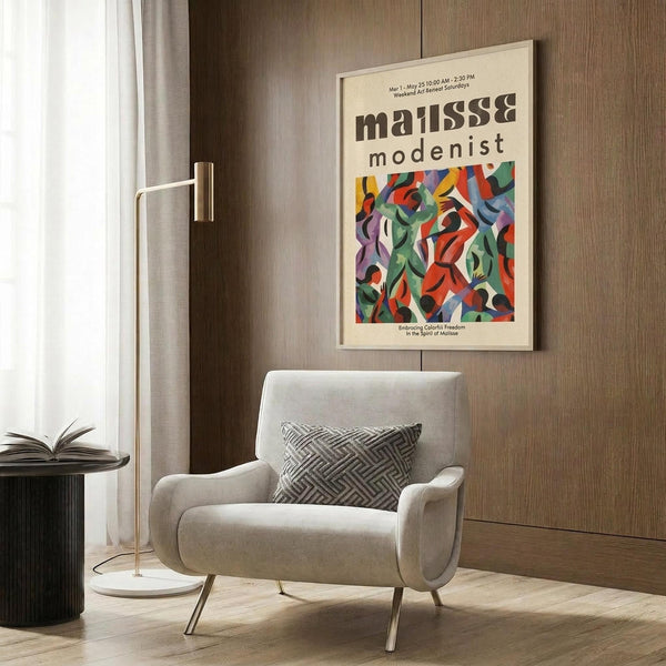 May include: Beige framed vintage-style exhibition poster featuring colorful abstract cut-out shapes in red, green, blue, and purple, displayed on wood-paneled wall above cream upholstered armchair with geometric patterned pillow, beside brass floor lamp and dark round side table with open book.