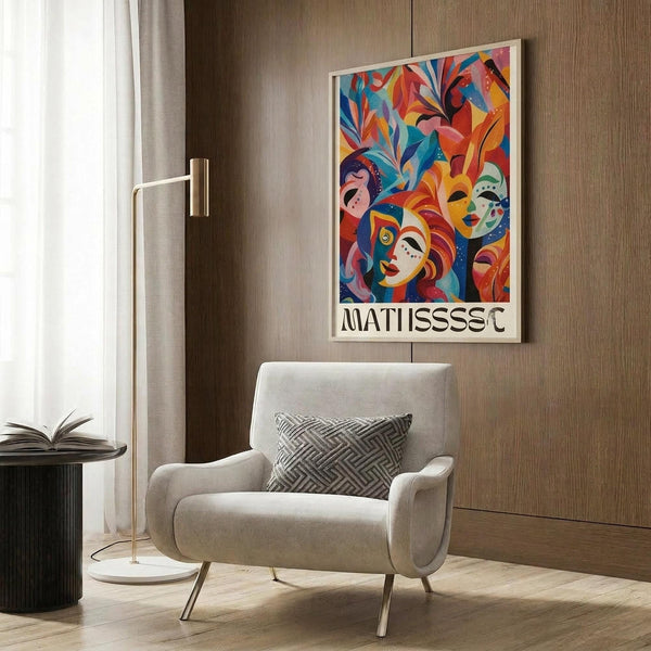 May include: Colorful abstract art poster featuring vibrant faces, masks, and foliage in red, blue, orange, and pink tones, displayed in light wood frame on wood-paneled wall above beige upholstered chair with geometric pillow, alongside brass floor lamp and dark round side table with open book
