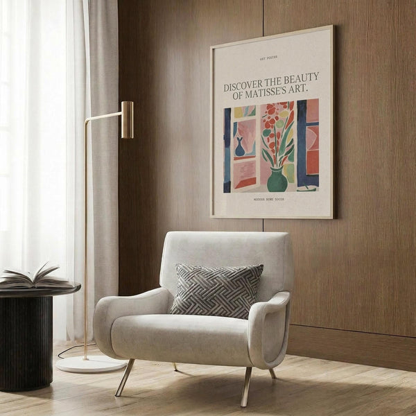 May include: Light gray upholstered armchair with geometric patterned pillow positioned next to wood-paneled wall. Framed colorful art print featuring abstract interior scene with flowers in green vase, displayed above chair. Gold floor lamp and dark round side table with open book complete the modern reading nook setting with natural lighting from nearby window.
