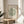 May include: Framed botanical art print in natural wood frame displayed on round wooden side table next to beige upholstered chair. Green coral-like shapes on cream background with "MATISSE" and "BERGGRUEN & CIE" text. Soft natural lighting creates shadows across white wall. Plant leaves visible in corner of minimalist room setting.