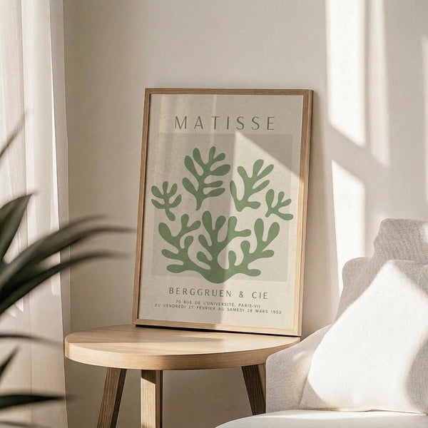 May include: Framed botanical art print in natural wood frame displayed on round wooden side table next to beige upholstered chair. Green coral-like shapes on cream background with "MATISSE" and "BERGGRUEN & CIE" text. Soft natural lighting creates shadows across white wall. Plant leaves visible in corner of minimalist room setting.