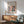May include: Framed art poster featuring colorful abstract interior scene with green vase of flowers, geometric shapes in coral, blue, and pink tones. Displayed above mid-century modern wooden credenza with white ceramic lamp. Sage green upholstered chair with wooden arms positioned nearby. Natural sunlight creates soft shadows on white wall.