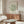 May include: Modern living room with cream walls featuring framed Matisse exhibition poster with green coral-like shapes on beige background. Warm wood sideboard below, white curved chair, glass oval coffee table, and minimalist lamp create mid-century inspired interior styling.