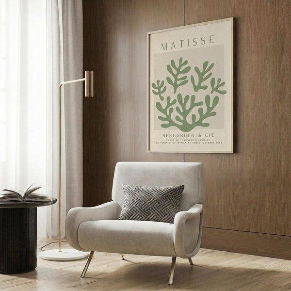 May include: Beige armchair with geometric patterned pillow in modern living room setting. Framed botanical poster with green leaf design and "MATISSE" text hangs on wood-paneled wall. Gold floor lamp, round black side table with open book, and natural lighting from window complete the warm, contemporary interior scene.