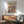 May include: Framed modernist art poster with colorful abstract figures in red, green, blue, and purple tones displayed above a wooden sideboard. Mid-century modern room setting features sage green upholstered chair with wood arms, white ceramic table lamp, and round wooden side table. Natural sunlight creates geometric shadows on white wall, highlighting the poster's vibrant cubist-style artwork and exhibition text.