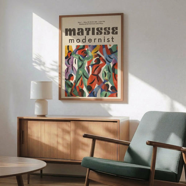 May include: Framed modernist art poster with colorful abstract figures in red, green, blue, and purple tones displayed above a wooden sideboard. Mid-century modern room setting features sage green upholstered chair with wood arms, white ceramic table lamp, and round wooden side table. Natural sunlight creates geometric shadows on white wall, highlighting the poster's vibrant cubist-style artwork and exhibition text.