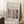 May include: Framed art poster leaning against white wall corner featuring colorful Matisse-style interior scene with green vase of red flowers, blue window frames, and geometric shapes in pink, coral, and yellow tones. White coffee cup on wooden block nearby, beige textured rug below, soft natural lighting creates shadows on wall.