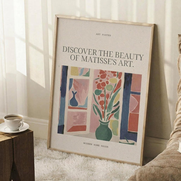 May include: Framed art poster leaning against white wall corner featuring colorful Matisse-style interior scene with green vase of red flowers, blue window frames, and geometric shapes in pink, coral, and yellow tones. White coffee cup on wooden block nearby, beige textured rug below, soft natural lighting creates shadows on wall.