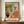 May include: Framed art print displayed on wooden sideboard featuring colorful interior scene with orange flowers in black vase, pink upholstered chair with leopard print pillow, and tropical plants. Warm peachy walls visible through doorway. Matisse text at bottom. White ceramic vase and books shown alongside for scale in neutral room setting.
