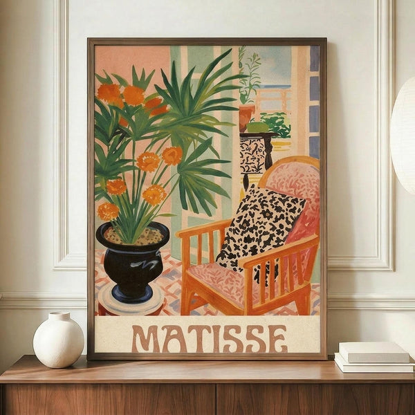 May include: Framed art print displayed on wooden sideboard featuring colorful interior scene with orange flowers in black vase, pink upholstered chair with leopard print pillow, and tropical plants. Warm peachy walls visible through doorway. Matisse text at bottom. White ceramic vase and books shown alongside for scale in neutral room setting.