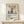 May include: Matisse-inspired poster in light wood frame leaning against white wall on wooden shelf, featuring colorful still life with blue vases, red fruits, yellow lemon, and botanical elements in cut-paper collage style. Small wrapped gift box with greenery sits in front. Warm natural lighting creates soft shadows on cream background.