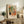 May include: Framed Matisse-style art print displayed on rustic wooden console table alongside fern plant, books, and ceramic vase. Print features colorful interior scene with orange flowers in black pot, person in patterned clothing sitting in orange chair, and window view. Warm lighting creates cozy living room atmosphere with neutral walls and brown leather furniture visible.