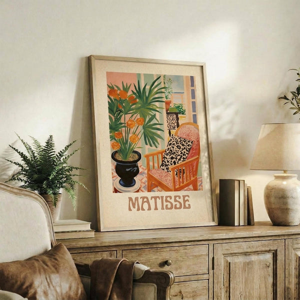 May include: Framed Matisse-style art print displayed on rustic wooden console table alongside fern plant, books, and ceramic vase. Print features colorful interior scene with orange flowers in black pot, person in patterned clothing sitting in orange chair, and window view. Warm lighting creates cozy living room atmosphere with neutral walls and brown leather furniture visible.