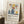 May include: Framed poster with Matisse-inspired still life artwork featuring blue ceramic vases, colorful fruit including yellow lemons and red oranges, green foliage, displayed on cream textured paper. Shows "Celebrating Colors of Life" text below illustration. Frame leans against white wall corner next to wooden side table with white coffee cup, cream knit throw blanket visible, creating cozy home interior mockup scene.