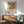 May include: Framed botanical art print with colorful flowers and leaves in Matisse-inspired style, displayed above wooden sideboard with ceramic lamp. Mid-century modern living room setting features sage green upholstered armchair with wooden frame and round coffee table. Natural sunlight creates shadows on white wall. Print shows vibrant orange, pink, and green botanical motifs with descriptive text below artwork.
