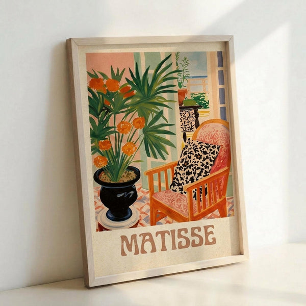 A framed art print in a light wood frame leaning against a white wall. The artwork depicts a bright interior scene with an orange wooden chair featuring a black and white patterned cushion, a large black planter with orange flowers and green palm-like foliage, and a window showing other plants in the background. The style uses bold, flat colors in oranges, greens, blacks and warm tones with "MATISSE" printed at the bottom in brown lettering.