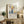 May include: Framed poster with Matisse-inspired still life artwork featuring colorful fruit arrangement in blue vases - red apples, yellow pear, green and blue foliage on cream background with "Celebrating Colors of Life" text below. Light wood frame displayed on rustic wooden sideboard alongside fern plant, books, and ceramic vase in neutral living room setting.