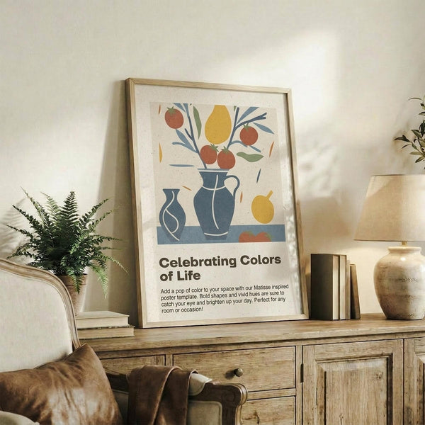 May include: Framed poster with Matisse-inspired still life artwork featuring colorful fruit arrangement in blue vases - red apples, yellow pear, green and blue foliage on cream background with "Celebrating Colors of Life" text below. Light wood frame displayed on rustic wooden sideboard alongside fern plant, books, and ceramic vase in neutral living room setting.