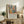 May include: Framed art print with colorful still life featuring yellow vase, green bottles, flowers, and fruit in modern abstract style, displayed on rustic wood sideboard next to fern plant and cream lamp in cozy living room setting with beige armchair and throw pillows