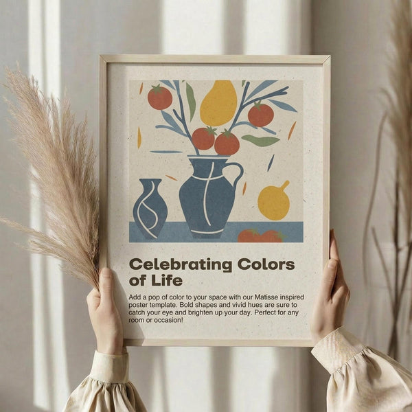 May include: Hands holding a framed art print featuring a colorful still life with blue pottery vases, red apples, yellow lemon, and green leaves in Matisse-inspired style. The artwork has bold, simplified shapes on cream textured paper with "Celebrating Colors of Life" text below. Natural wood frame, displayed against white curtained window with dried wheat stalks visible on left side.