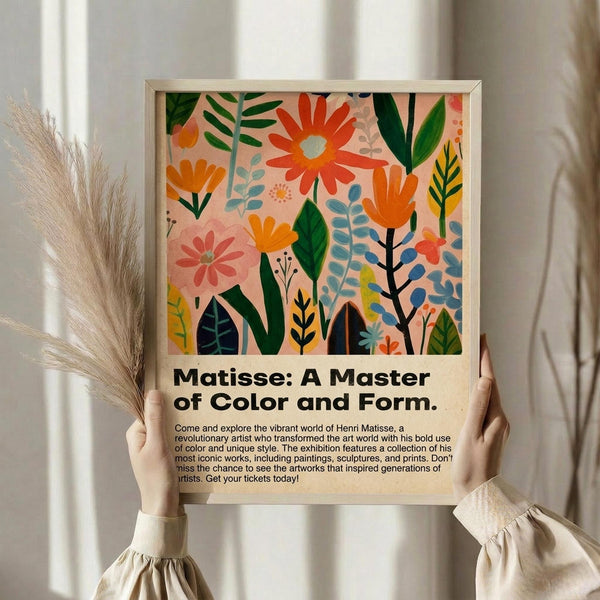 May include: Hands holding a framed poster featuring colorful botanical artwork with vibrant flowers and leaves in orange, pink, green, and blue against a peachy background. Below the artwork is text about a Matisse exhibition. The wooden frame is displayed against white curtains with dried pampas grass visible to the left. The person wears a cream-colored blouse with ruffled sleeves.
