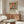 May include: Colorful botanical poster with vibrant flowers and leaves in orange, pink, green, and blue tones, displayed in thin wooden frame above walnut credenza. Modern living room setting features white sculptural chair, glass-top oval coffee table, and natural lighting creating soft shadows on cream walls.