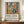 May include: Framed exhibition poster featuring colorful still life artwork with vases, flowers, and fruit in Matisse-inspired style, displayed on wooden sideboard next to white ceramic vase and books against cream wainscoted wall. Poster shows vibrant yellows, greens, oranges, and blues with bold geometric shapes and floral motifs. Brown wooden frame complements warm interior setting.