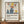 May include: Framed poster with Matisse-inspired still life artwork featuring blue ceramic vases, colorful fruit including red apples and yellow pear, green foliage on cream textured background. Text reads "Celebrating Colors of Life" with description below. Brown wooden frame displayed on wood sideboard next to white ceramic vase and stacked books in neutral-toned room with white paneled walls.