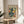 May include: Framed art poster displaying abstract blue female figure in Matisse's cut-out style with yellow and orange geometric shapes on cream background, positioned on round wooden side table next to beige upholstered chair, with plant leaves visible and natural sunlight creating shadows on white wall.