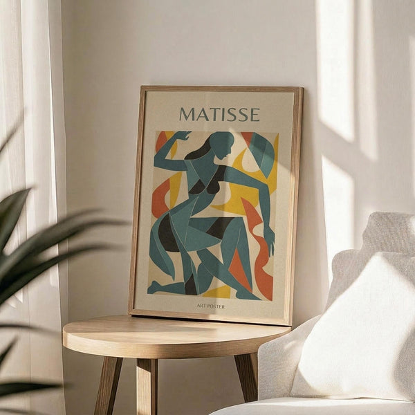 May include: Framed art poster displaying abstract blue female figure in Matisse's cut-out style with yellow and orange geometric shapes on cream background, positioned on round wooden side table next to beige upholstered chair, with plant leaves visible and natural sunlight creating shadows on white wall.