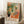 May include: Framed art print leaning against white wall corner showing colorful interior scene with orange wooden chair, pink cushion, black and white patterned throw pillow, dark vase with orange flowers and green palm fronds, patterned floor tiles, and window view. Artist name "MATISSE" appears at bottom. Coffee cup on wooden block and beige throw blanket visible in cozy room setting.