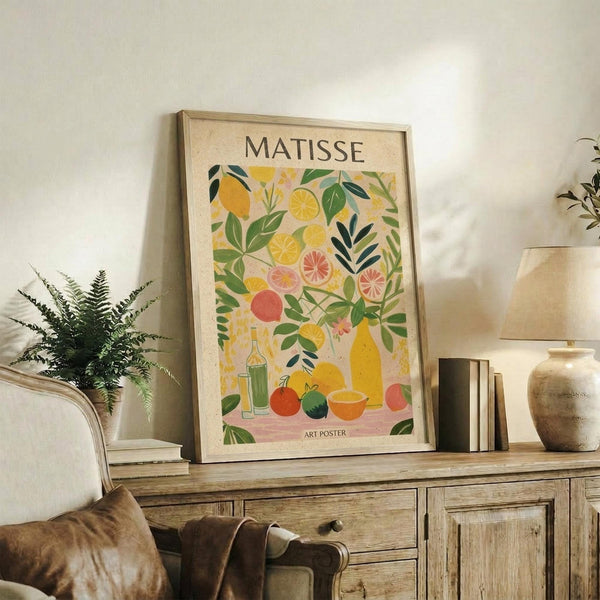 May include: Framed art poster with colorful fruit and botanical illustration in Matisse-inspired style, featuring yellow lemons, pink grapefruit, green leaves, and bottles on cream background. Displayed on rustic wooden sideboard with fern plant, books, ceramic vase, and brown leather chair in warm-toned living room setting.