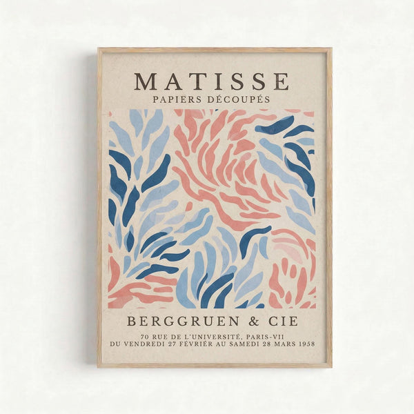 Framed vintage exhibition poster featuring abstract cut-paper designs in coral pink and blue tones on cream background. The artwork displays flowing, organic shapes resembling seaweed or flames. Text indicates a 1958 Paris gallery show. Natural wood frame against white wall in minimalist interior setting.