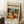 May include: Framed poster displaying a vibrant still life scene with wine bottle, glass, colorful fruits including oranges and grapes, leafy plants, and decorative bowls in Matisse-inspired bold colors. The cream-colored poster shows exhibition text at top and bottom. Frame leans against white wall corner next to wooden side table with white coffee cup, positioned on textured cream rug beside beige upholstered furniture.
