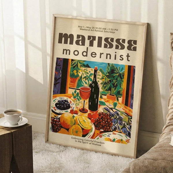 May include: Framed poster displaying a vibrant still life scene with wine bottle, glass, colorful fruits including oranges and grapes, leafy plants, and decorative bowls in Matisse-inspired bold colors. The cream-colored poster shows exhibition text at top and bottom. Frame leans against white wall corner next to wooden side table with white coffee cup, positioned on textured cream rug beside beige upholstered furniture.