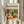 May include: Person holding framed modernist still life poster in light wood frame against white curtained window. Artwork shows vibrant fruits, wine bottle, and foliage in bold colors - oranges, greens, purples, reds. Dried pampas grass visible beside figure wearing cream colored ruffled sleeves.