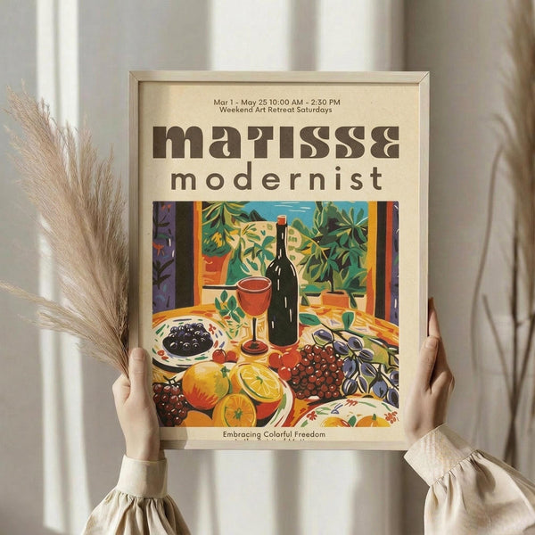 May include: Person holding framed modernist still life poster in light wood frame against white curtained window. Artwork shows vibrant fruits, wine bottle, and foliage in bold colors - oranges, greens, purples, reds. Dried pampas grass visible beside figure wearing cream colored ruffled sleeves.