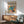 May include: Colorful modernist botanical poster with vibrant cut-out style leaves in teal, orange, pink and yellow, displayed in wooden frame above natural wood sideboard. Mid-century modern room setting features sage green upholstered chair with wooden arms, white ceramic table lamp, and warm sunlight casting geometric shadows on white wall.